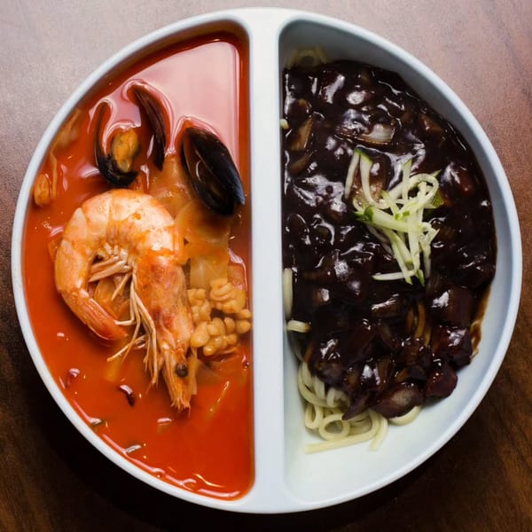 A divided bowl with red seafood soup on one side and black bean noodles on the other.